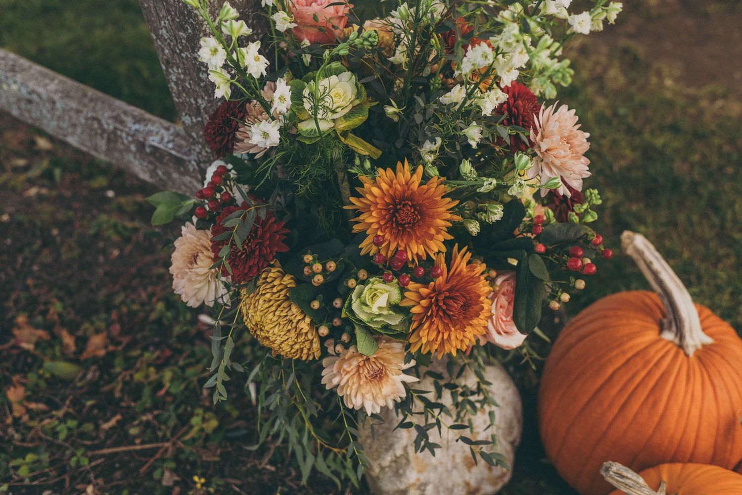 An autumn floral arrangement with sunflowers and dahlias in a rustic vase sits beside an orange pumpkin outdoors.