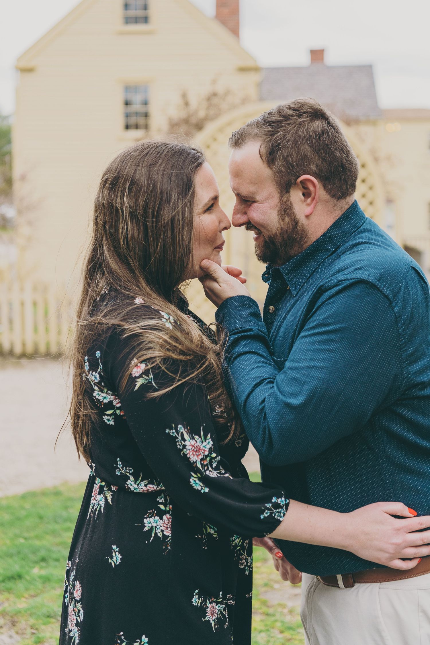 A romantic couple in blue and floral print clothing embracing outdoors against a beige house backdrop.