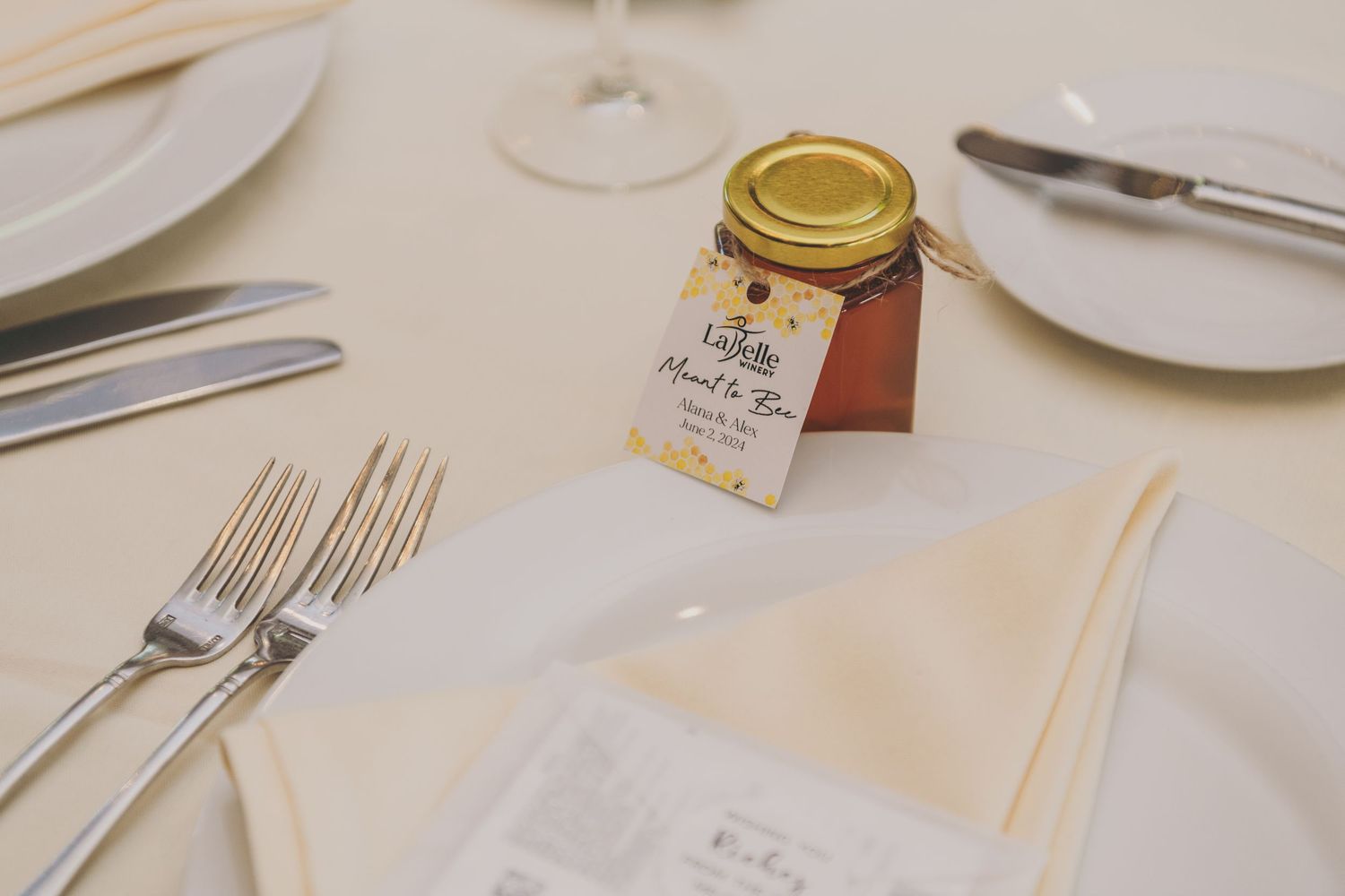 Wedding reception place setting with silverware, plates, and a small honey jar favor on a white tablecloth.