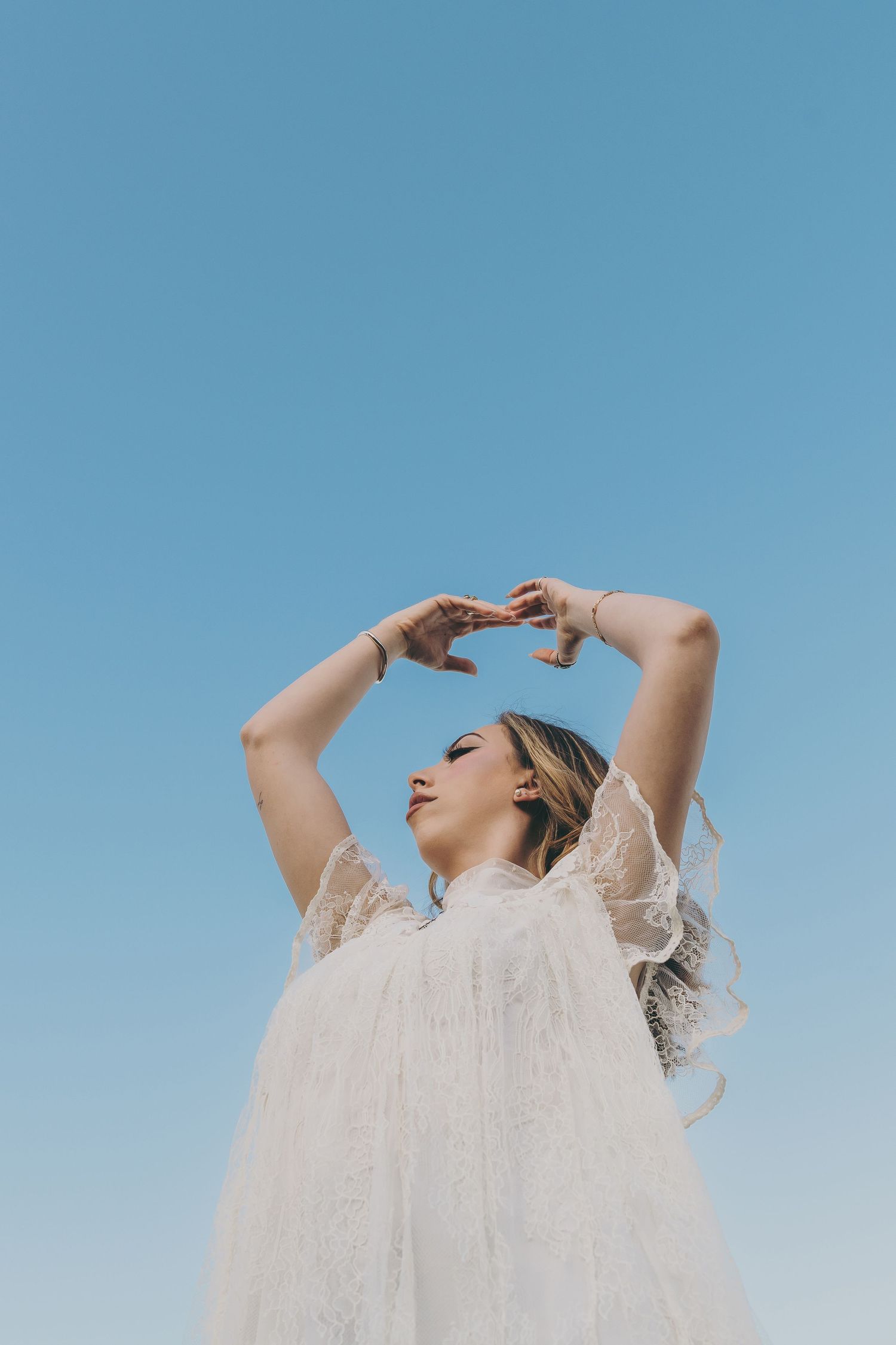 Figure in white lace dress poses against bright blue sky with arms raised.