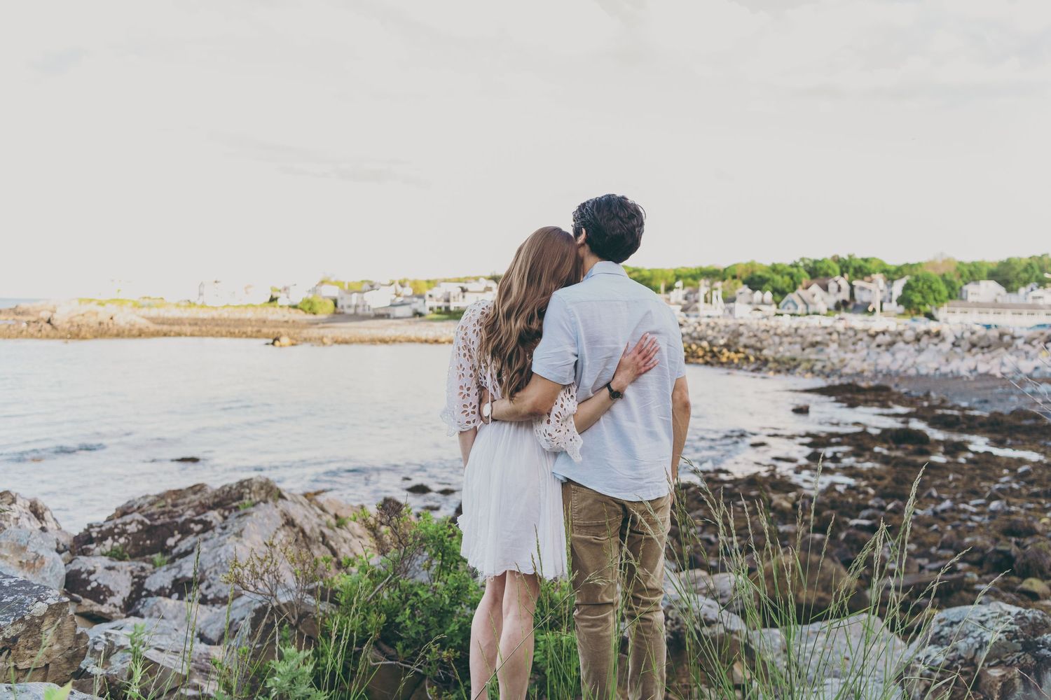 Couple embracing while standing on rocky coastal shore with ocean and beach houses in background during sunset.