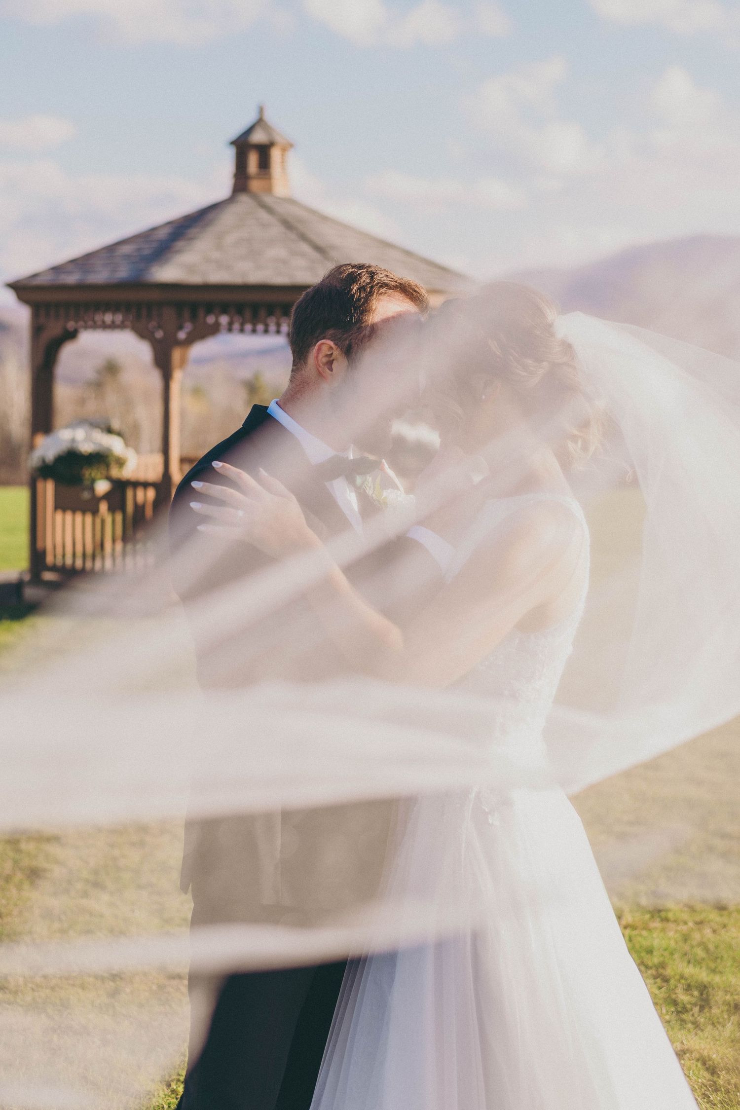 Wedding couple embraces outdoors as a flowing white veil billows in the breeze near a wooden gazebo.