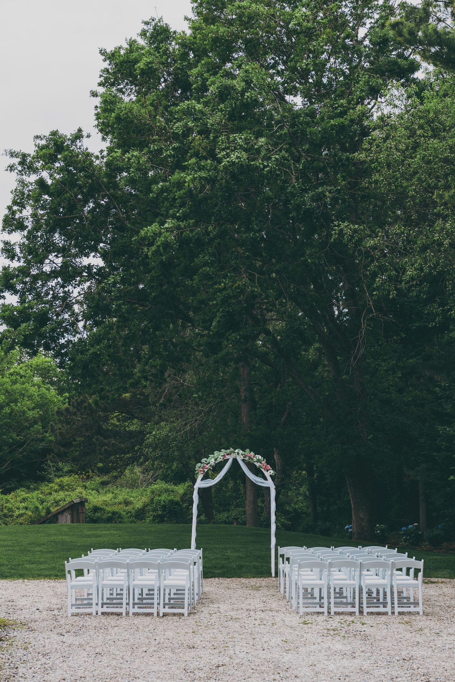 White wedding chairs arranged in rows for an outdoor ceremony facing a wooded backdrop on gravel ground.