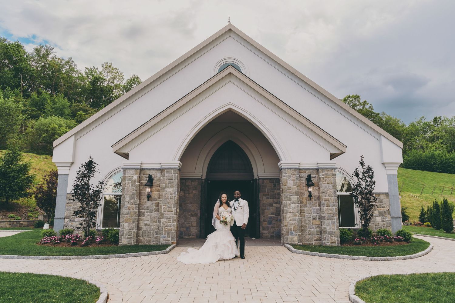 A couple stands in the entrance of a white stone chapel with an arched doorway and stone pillars on a cloudy day.