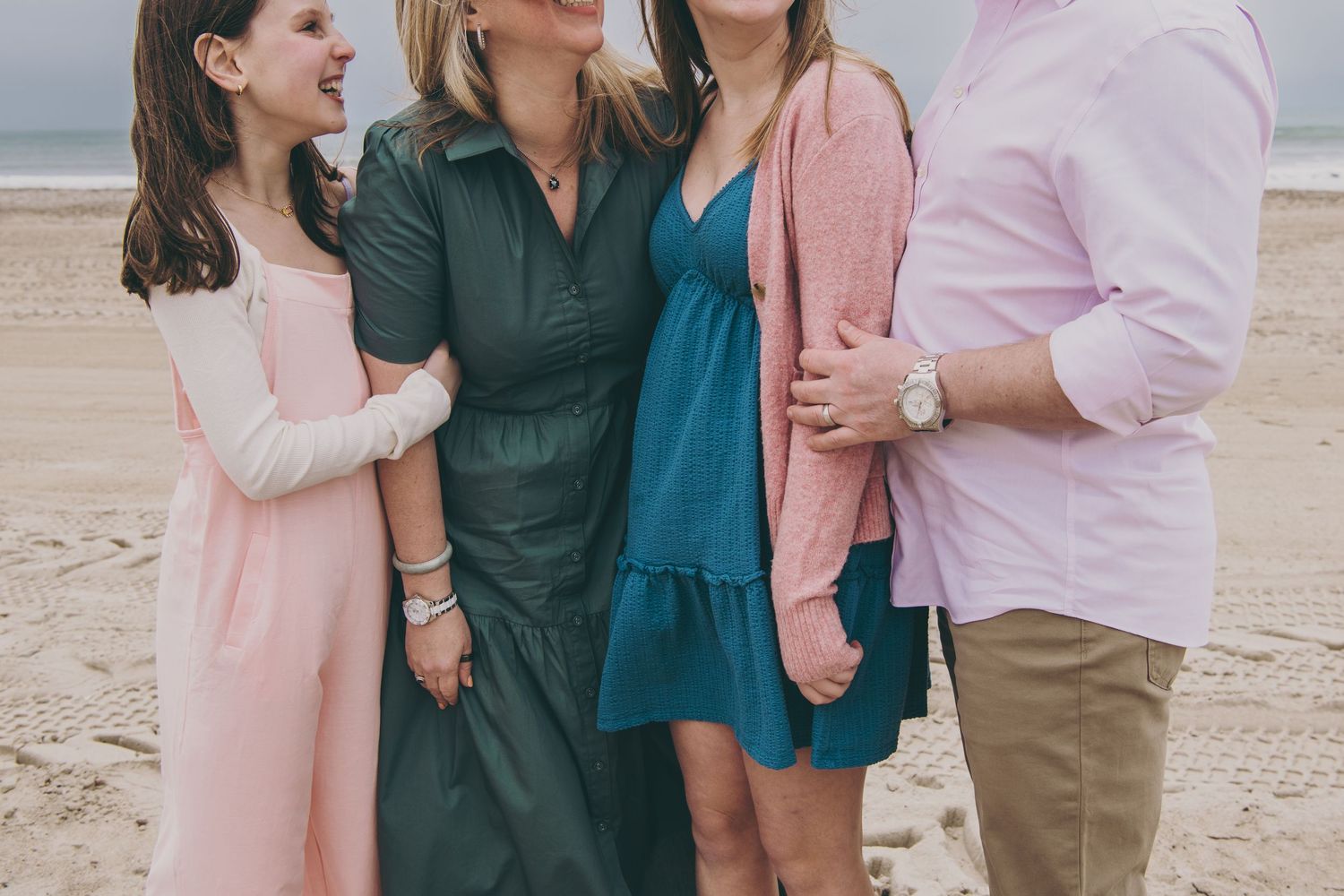A family wearing coordinated teal, pink and khaki outfits poses together for photos on the beach at sunset.
