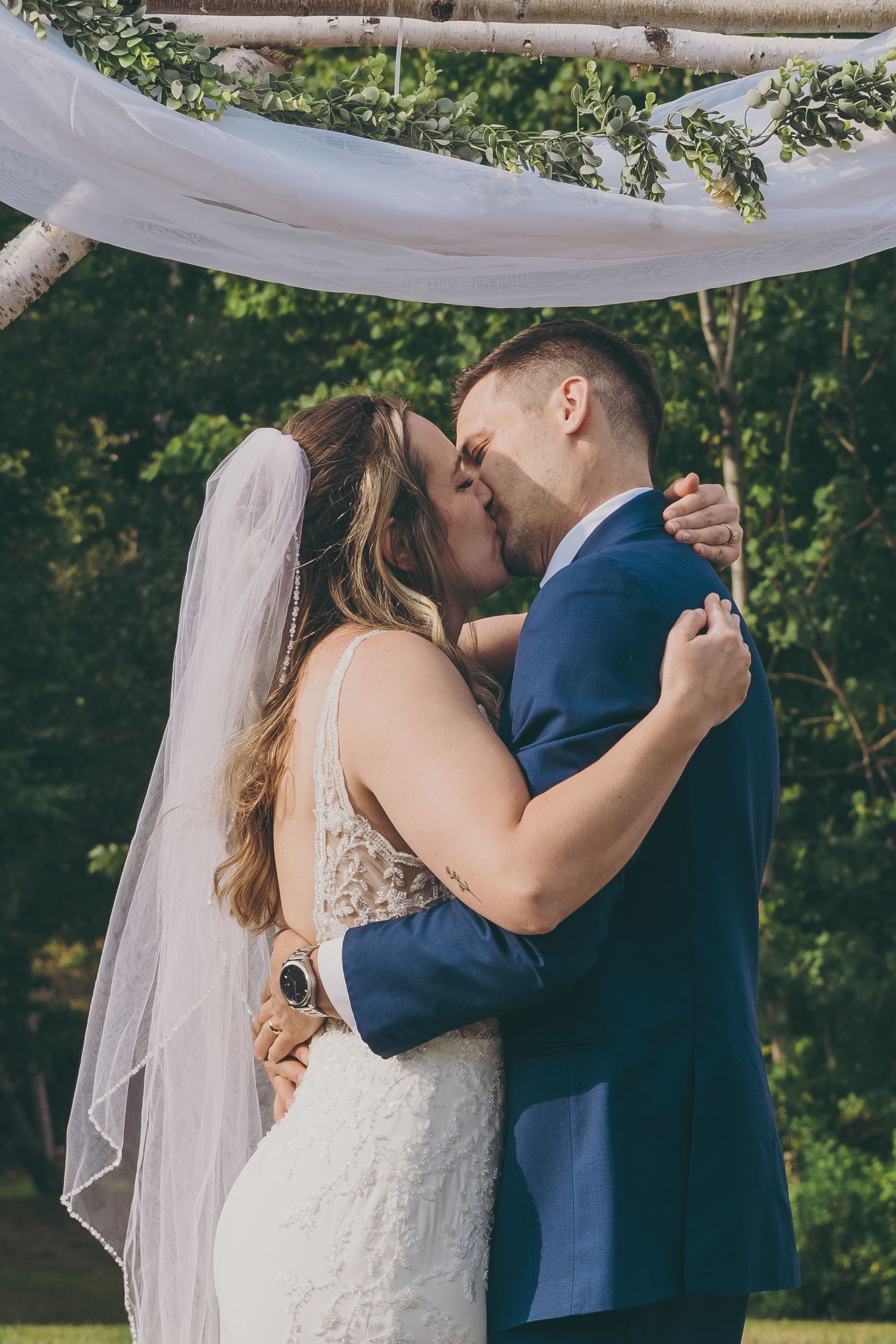 A romantic wedding couple shares their first kiss beneath a flowing white fabric canopy outdoors during their ceremony.