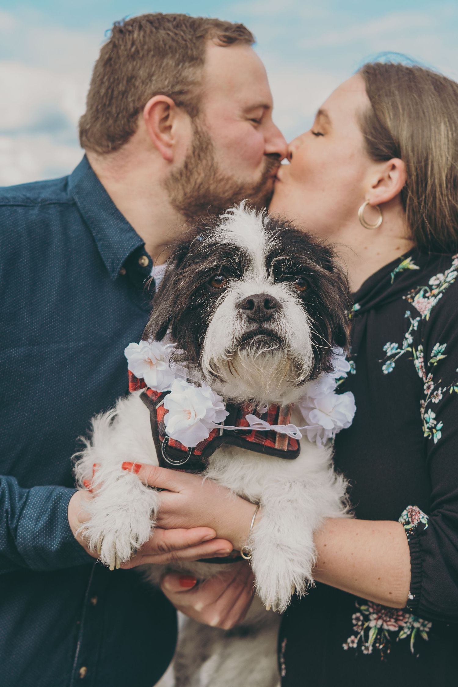 A couple shares a romantic kiss while holding their small dog wearing a bowtie.