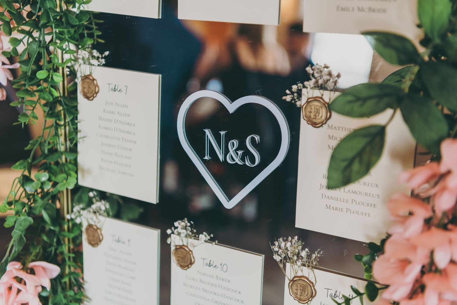 Wedding seating chart display with N&S heart logo surrounded by greenery and floral arrangements.