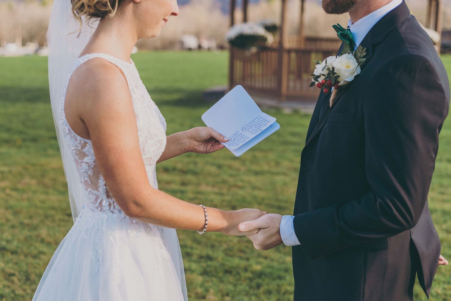 Couple holds hands during outdoor wedding ceremony as bride reads from blue paper, wearing white lace gown.