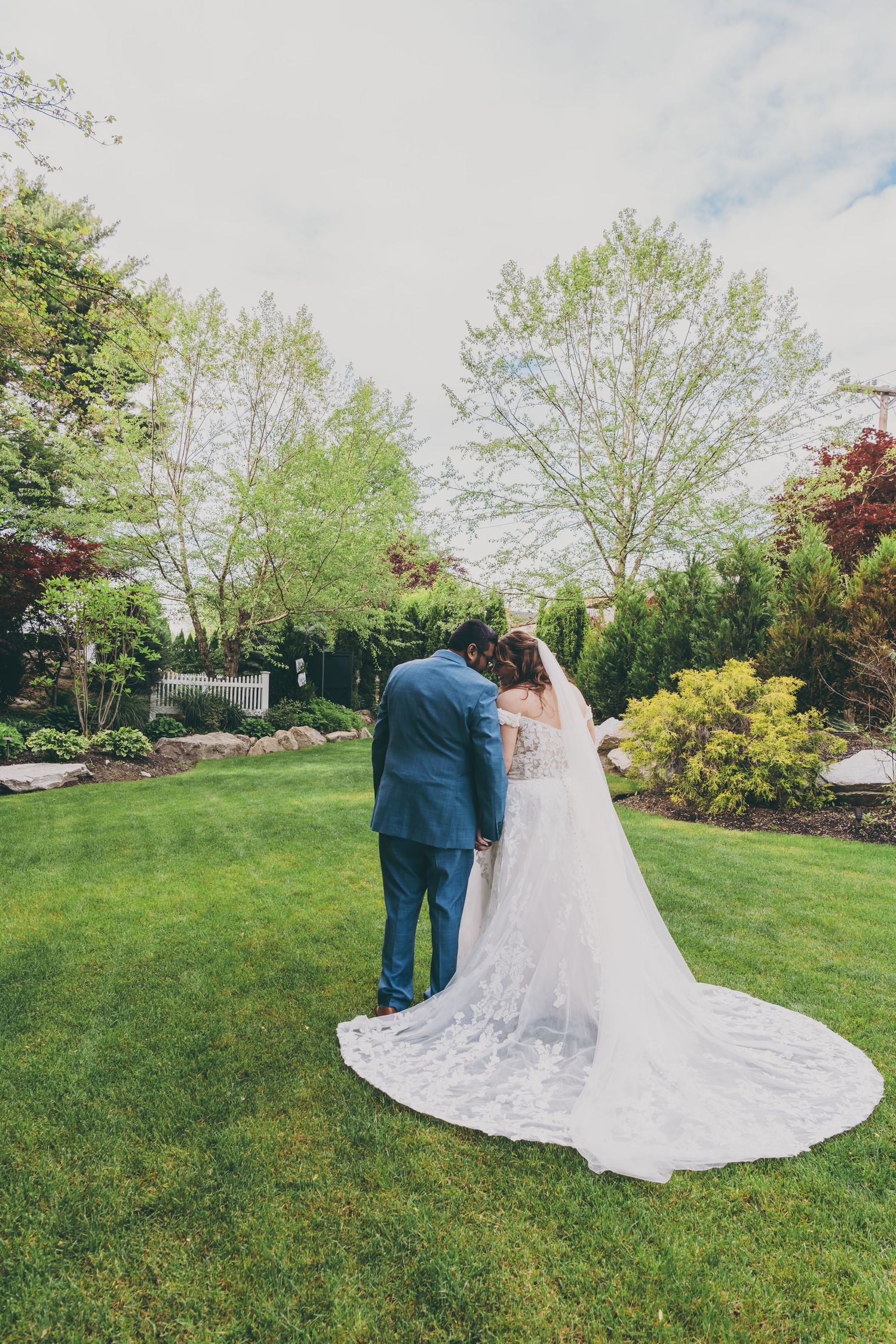 Couple embraces on manicured lawn with long lace wedding veil trailing behind in garden setting during spring.