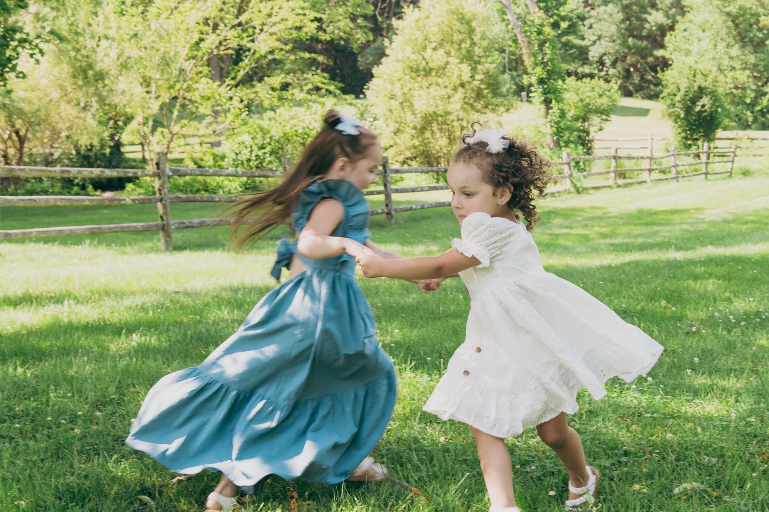 Two young children in dresses twirl and dance together in a sunny green park.