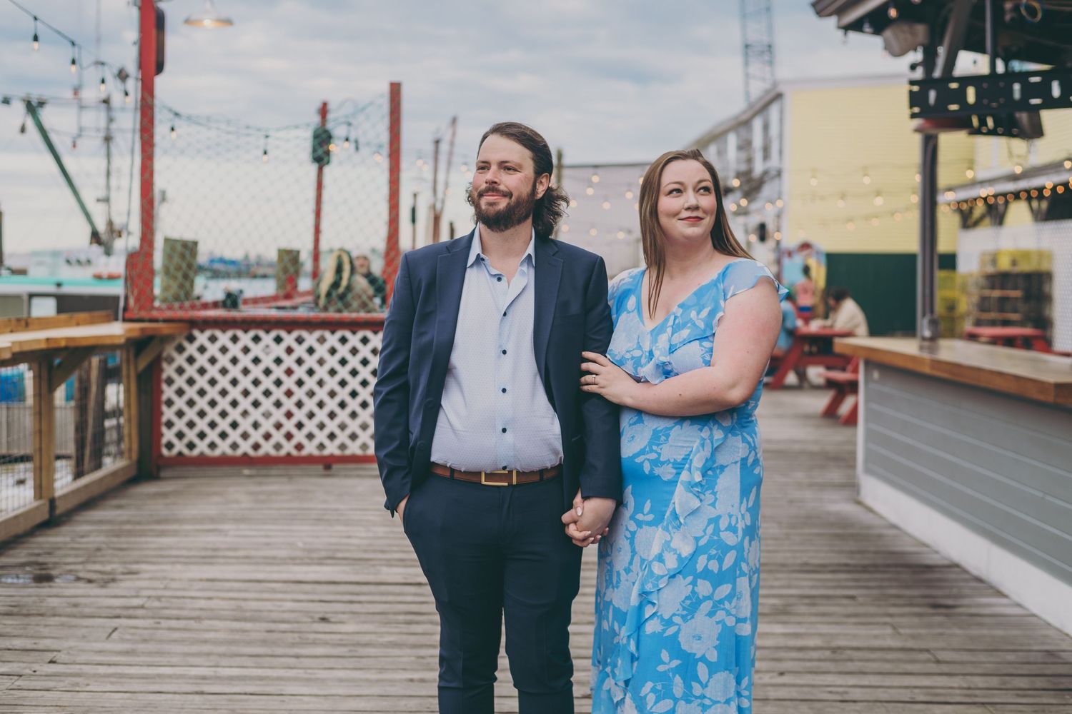 A couple standing on a wooden boardwalk with string lights and waterfront views.