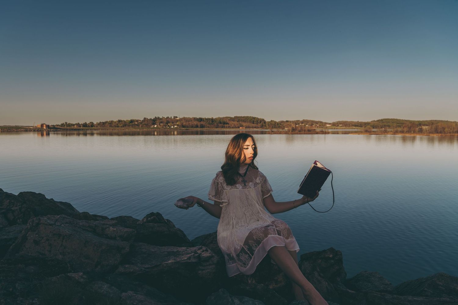 Someone in flowing dress sits on rocky shore with laptop against serene lake backdrop.