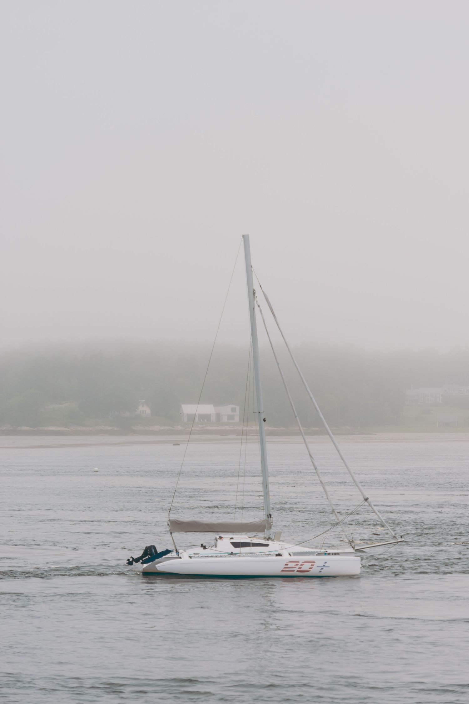 A white sailboat anchored in misty waters on a foggy day with distant shoreline barely visible in the background.