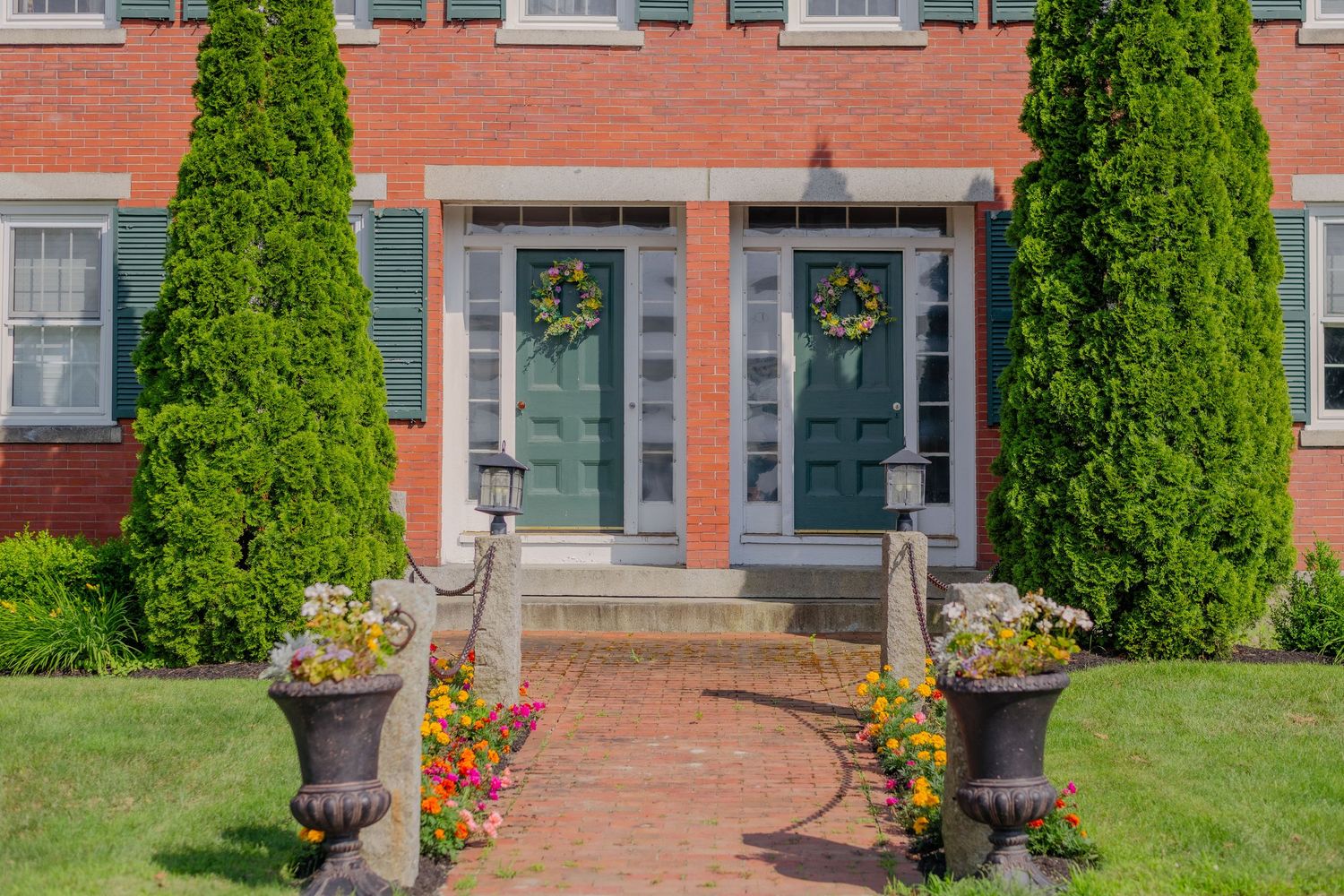 Historic red brick building with tall green shrubs and colorful flower planters along brick walkway in summer.