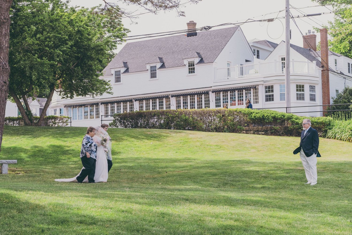 A white colonial style house with a covered porch sits on a manicured lawn where several people gather outdoors.
