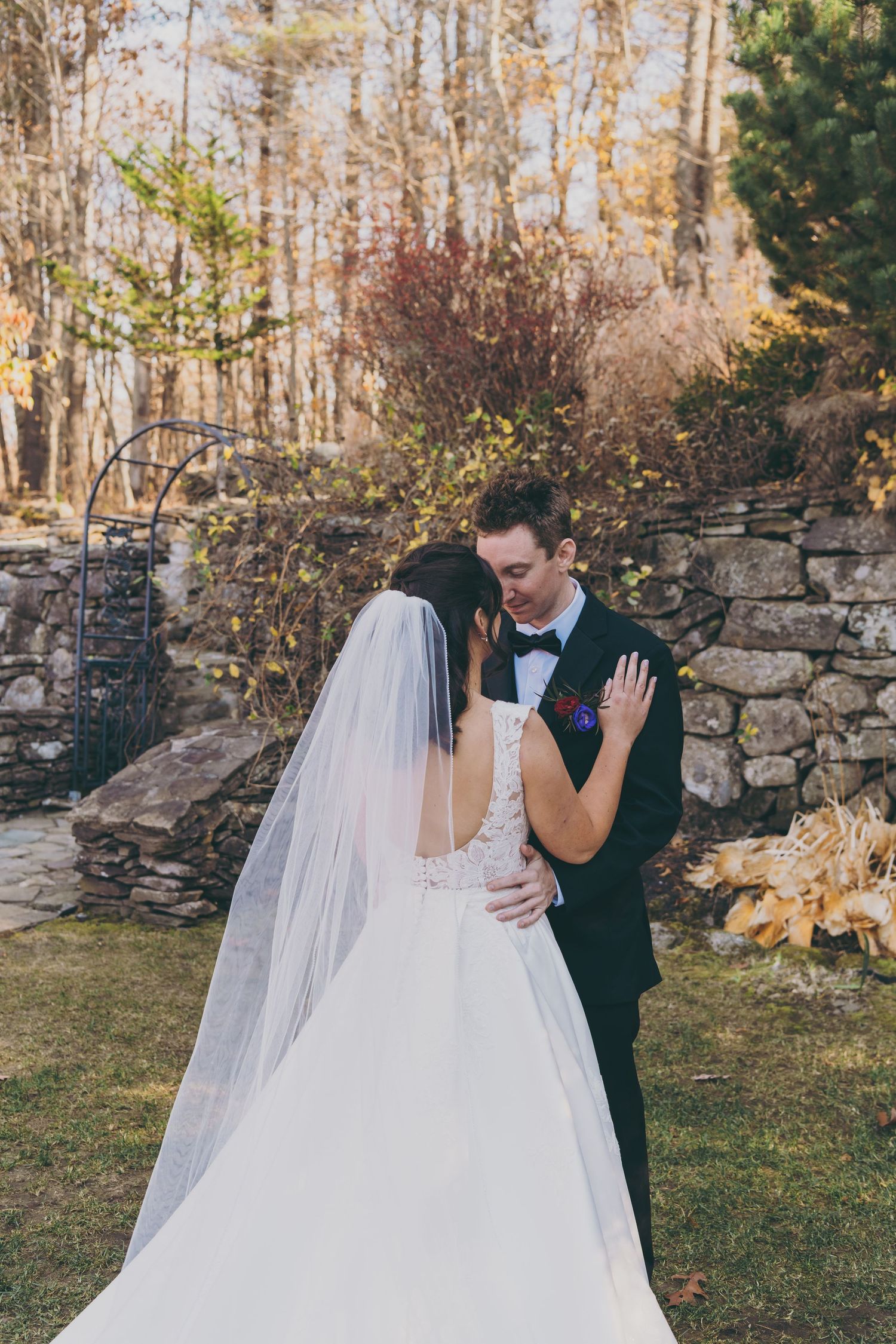 A couple in wedding attire embrace in front of a rustic stone wall during an autumn outdoor ceremony.