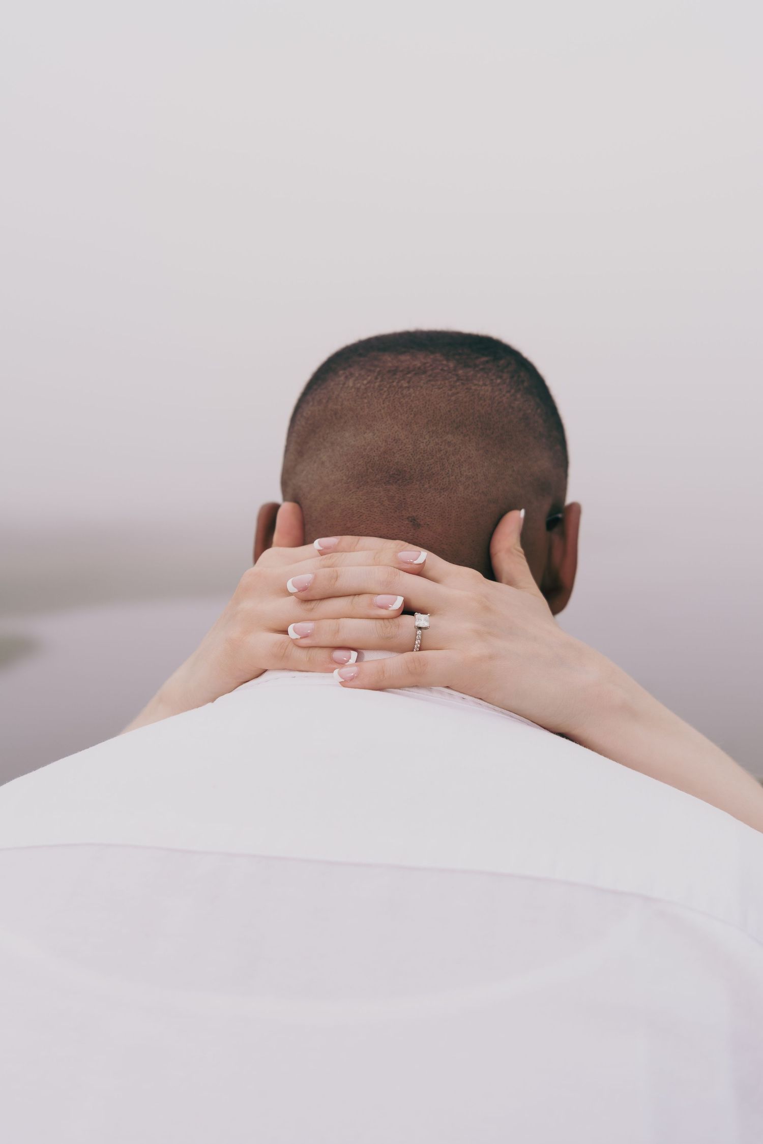 Hands with a ring gently resting on the back of someone's neck against a white shirt and light background.