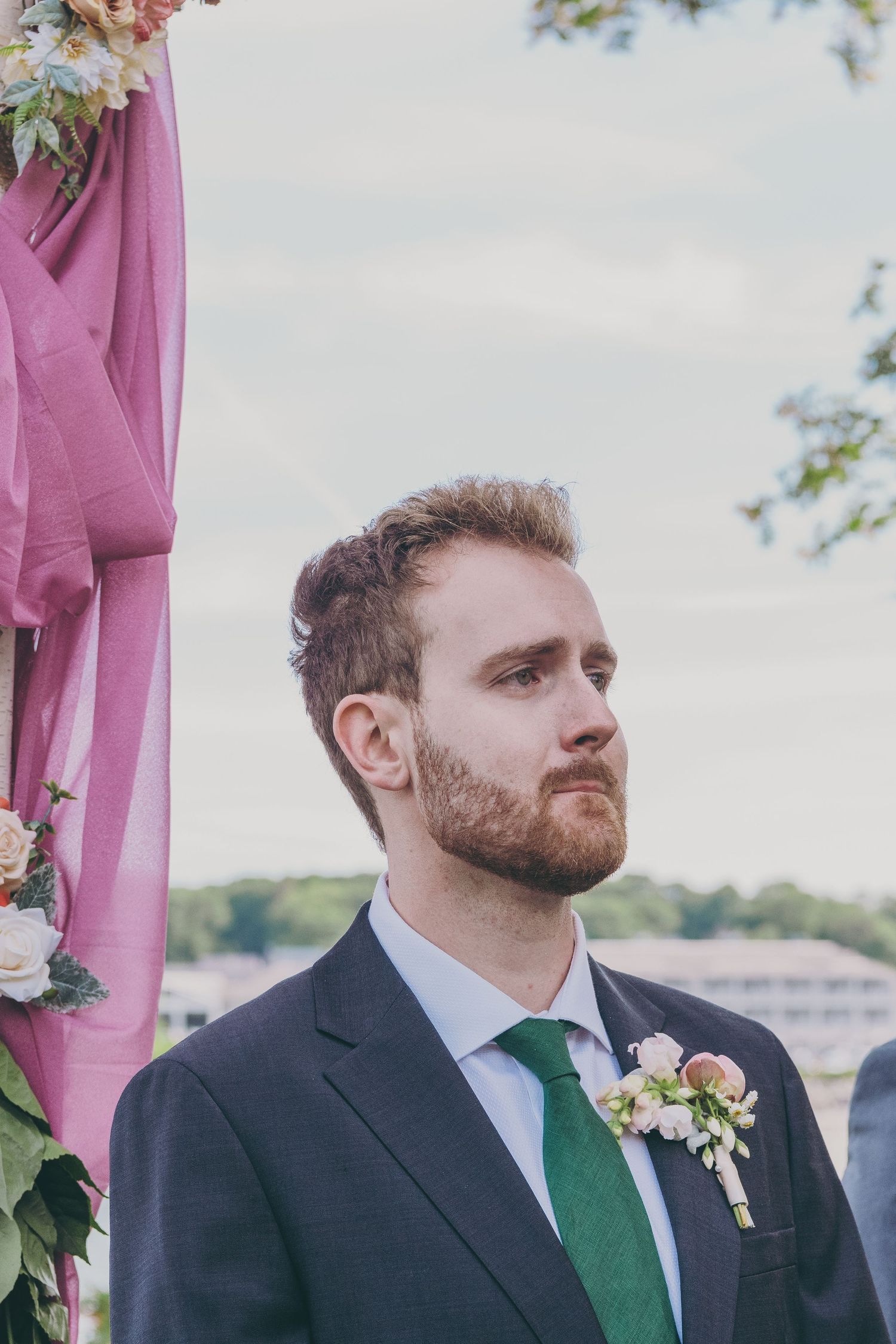 Wedding ceremony participant in dark suit and green tie stands beside pink fabric decor.