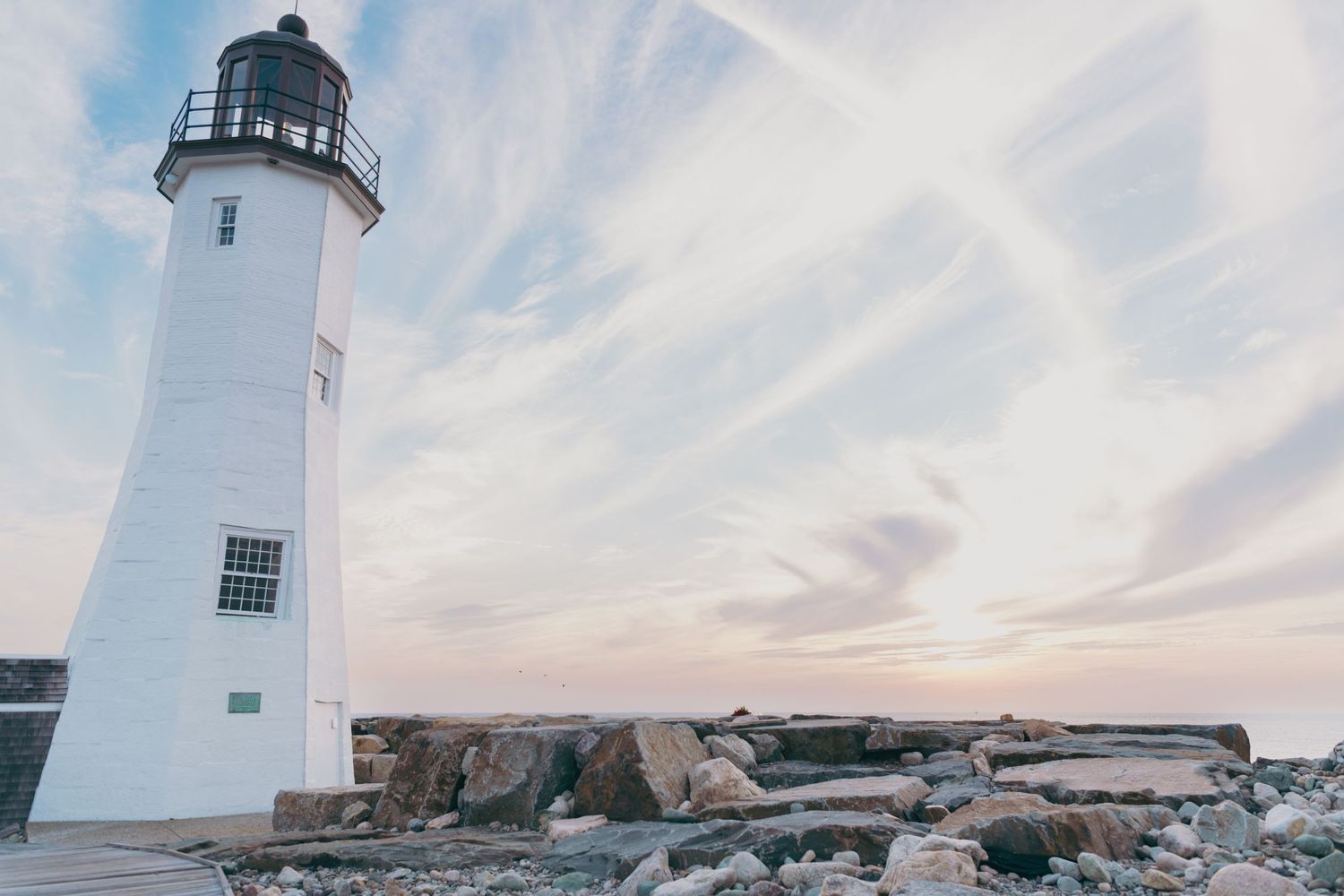 A white lighthouse stands tall on rocky shoreline against pastel sunset sky with wispy clouds. Scituate Lighthouse, Massachusetts
