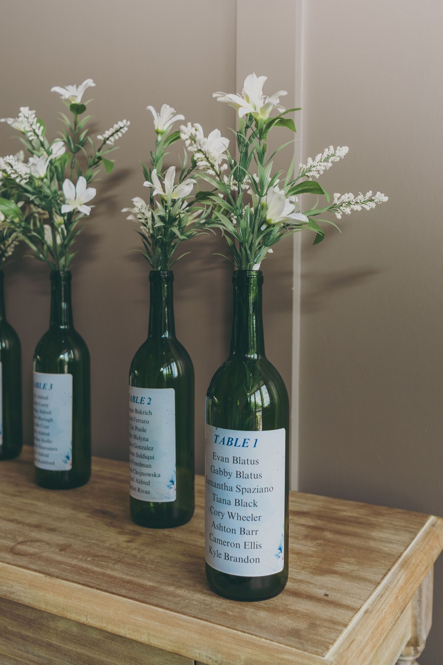 Row of green glass bottles with white flowers used as table number markers on wooden surface.