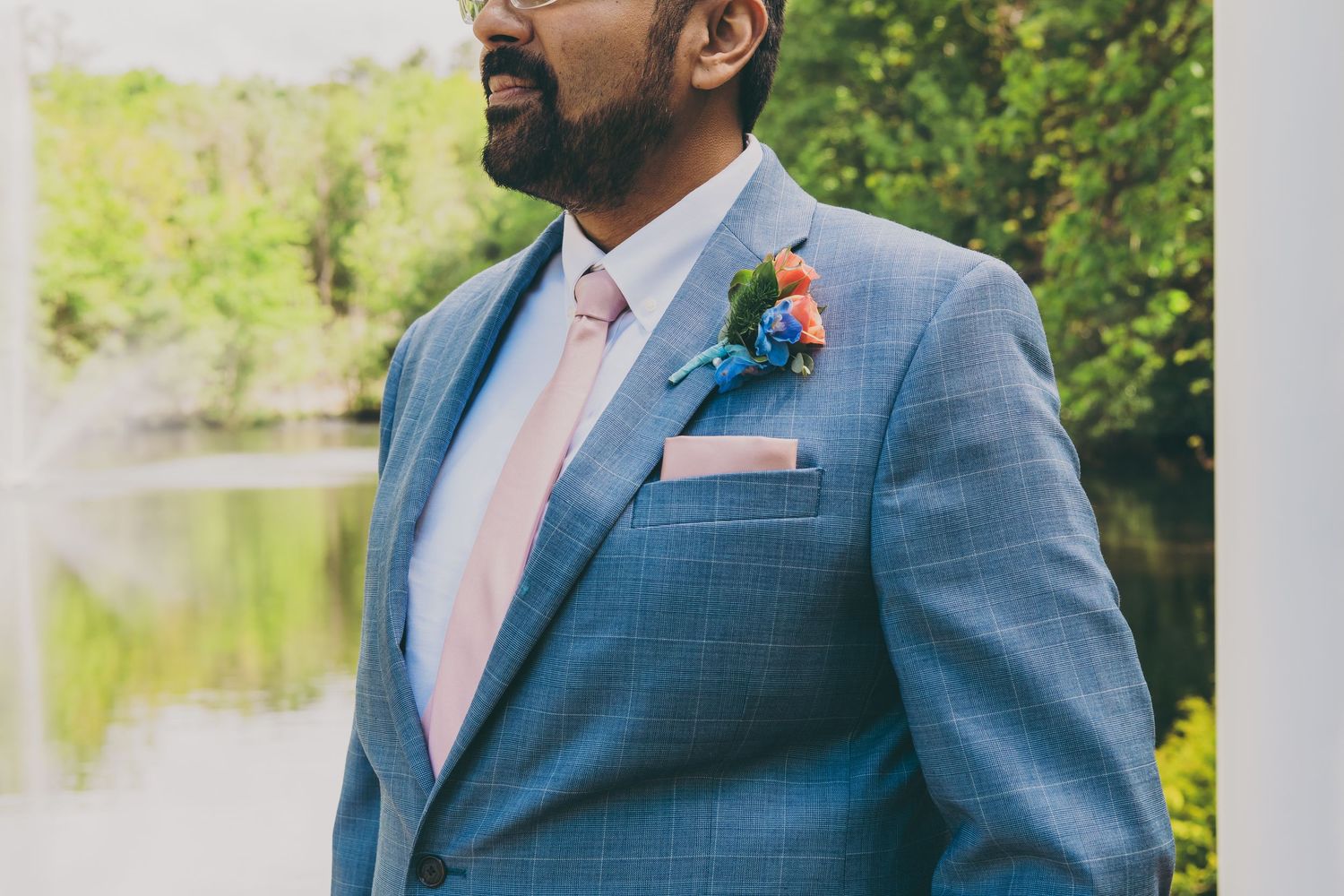 Wedding attire featuring a light blue suit with pink tie and coral boutonniere against a blurred outdoor background.