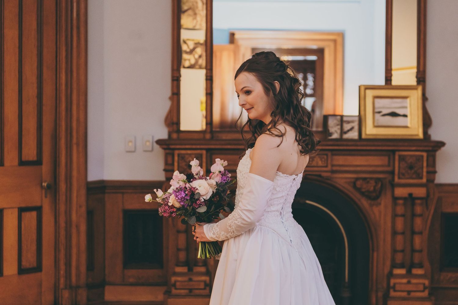 A bride in a flowing white gown holds a pink and white bouquet while standing in an elegant room.