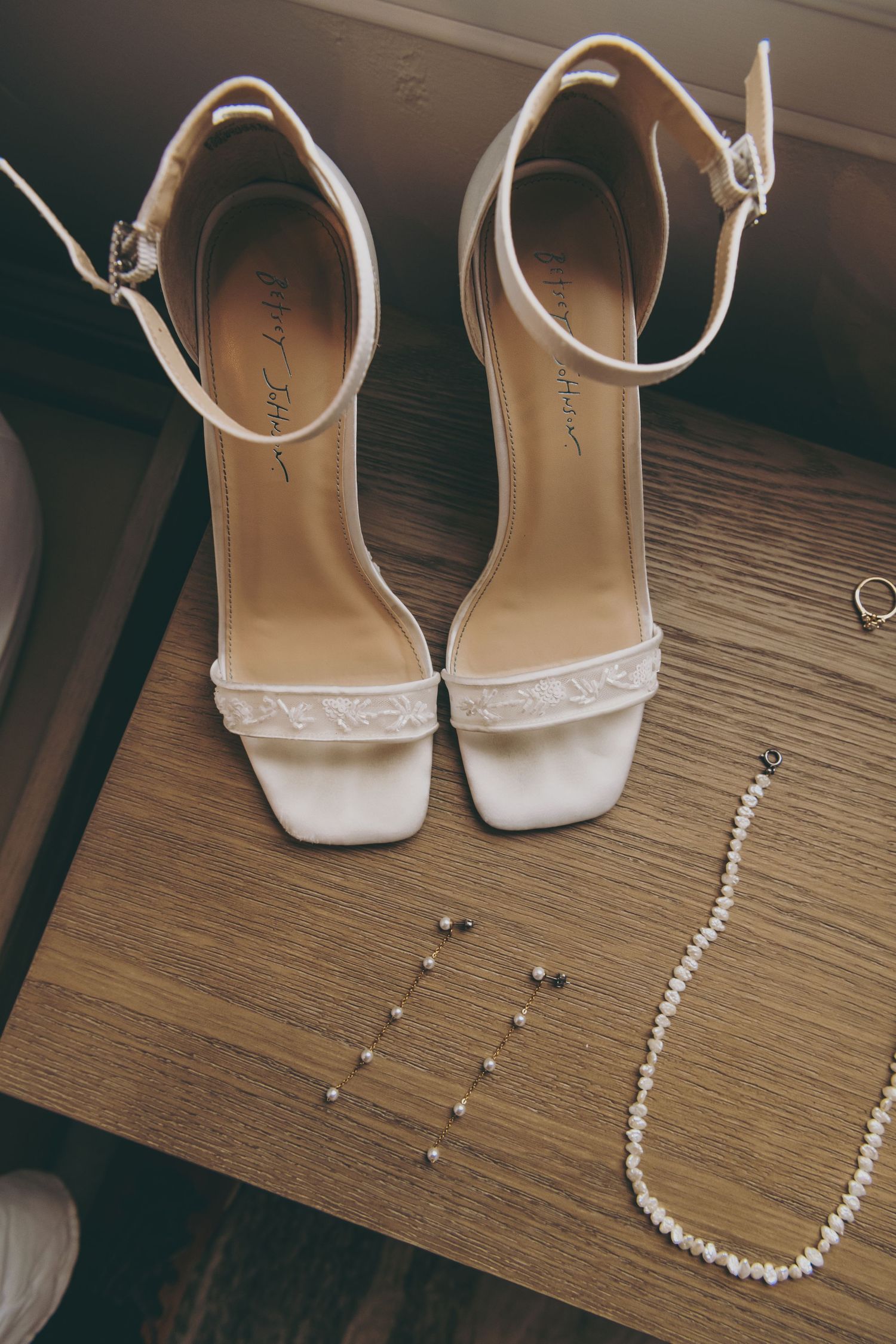 White bridal heels with delicate ankle straps displayed next to a pearl necklace and small earrings on wooden surface.