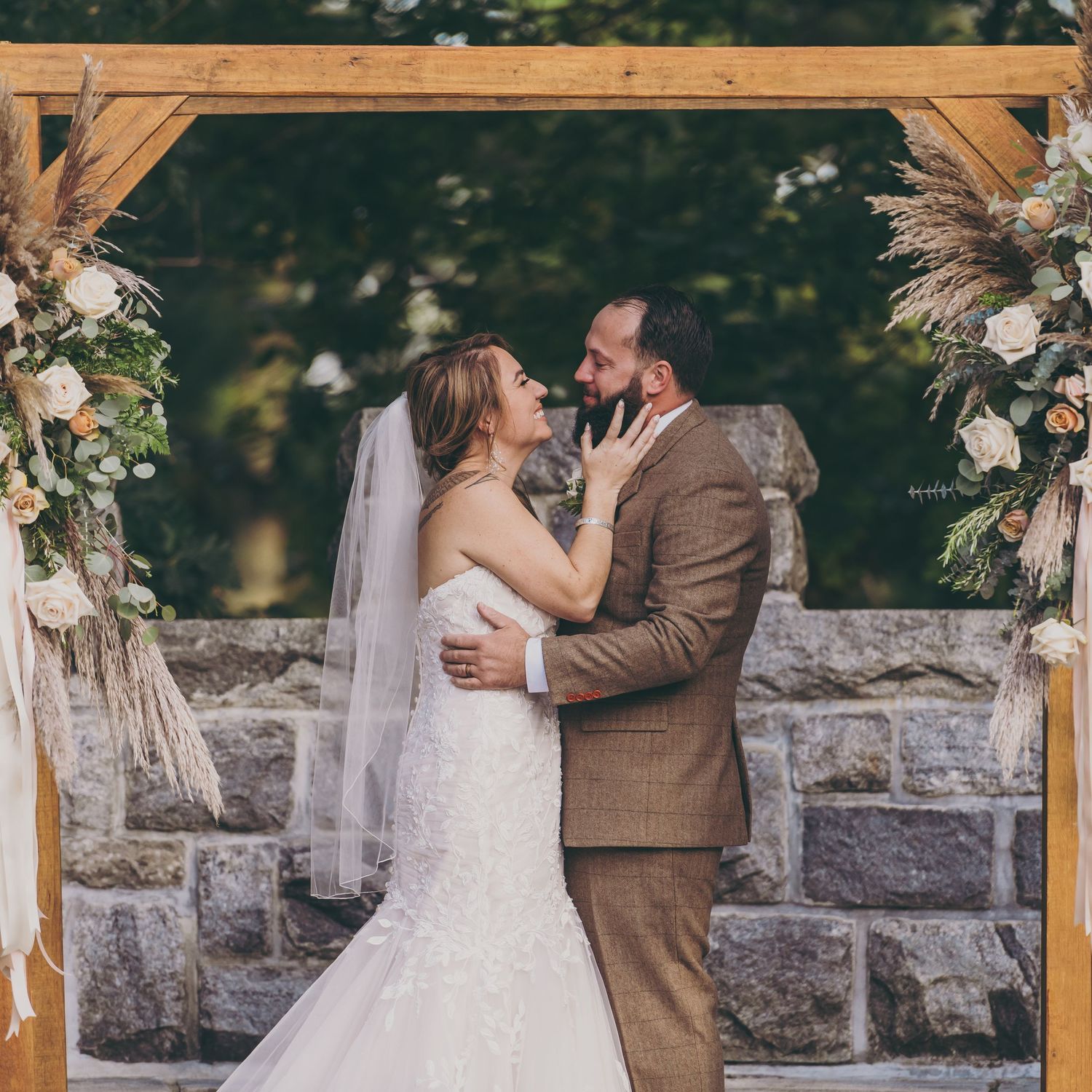 A romantic wedding ceremony under a wooden arch decorated with pampas grass and floral arrangements on a stone wall.
