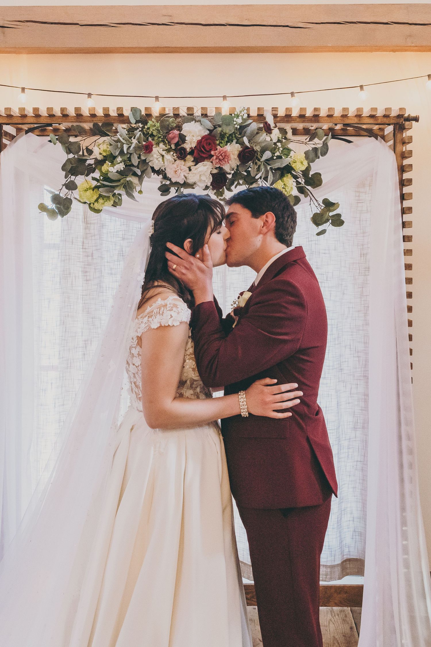 A romantic wedding moment under a floral arch with white drapes, featuring burgundy suit and white lace gown.