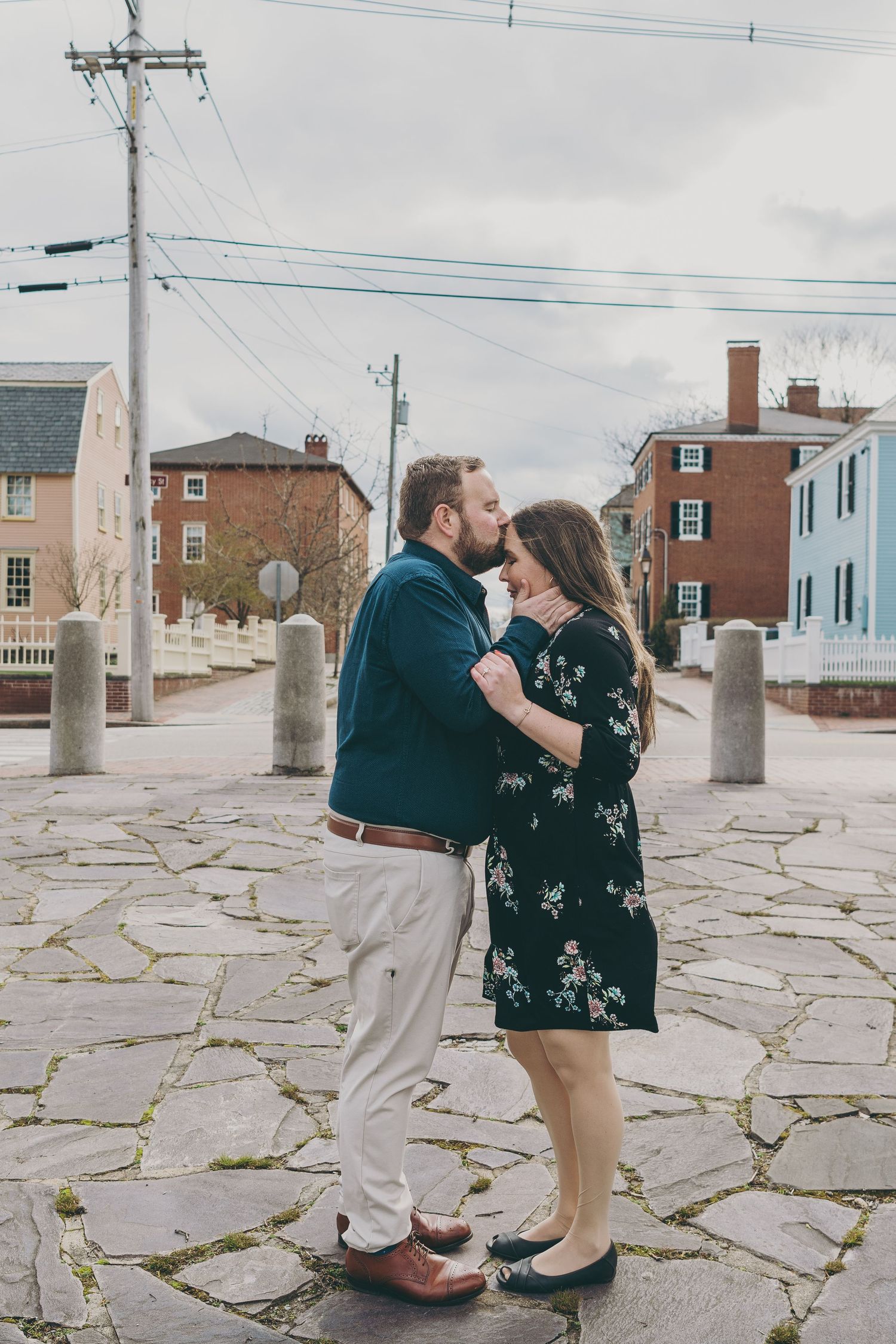 Two people share a romantic moment on a stone paved path with buildings and power lines in the background.