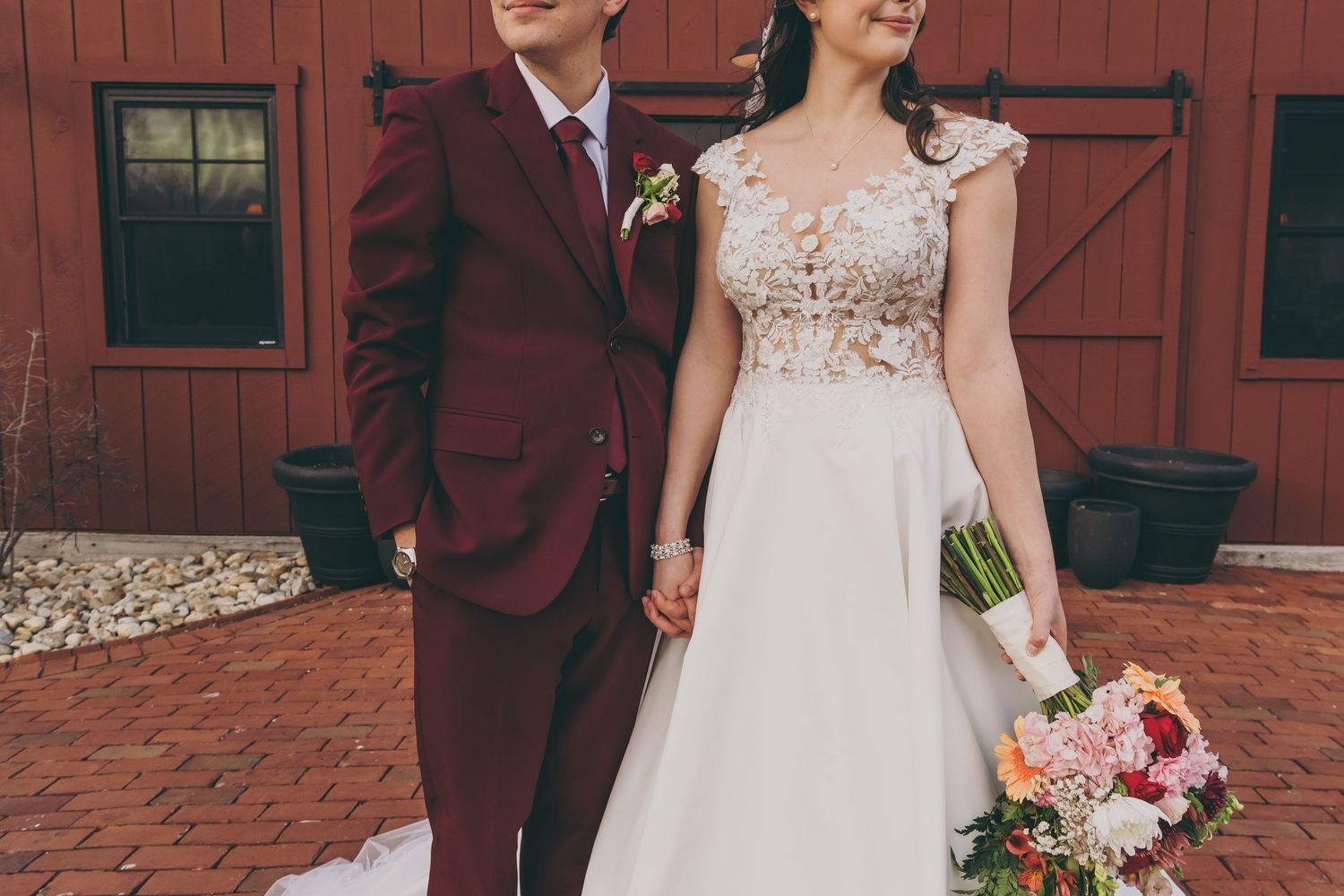 Wedding couple poses in front of a rustic red barn, the groom in burgundy and bride in white lace dress.