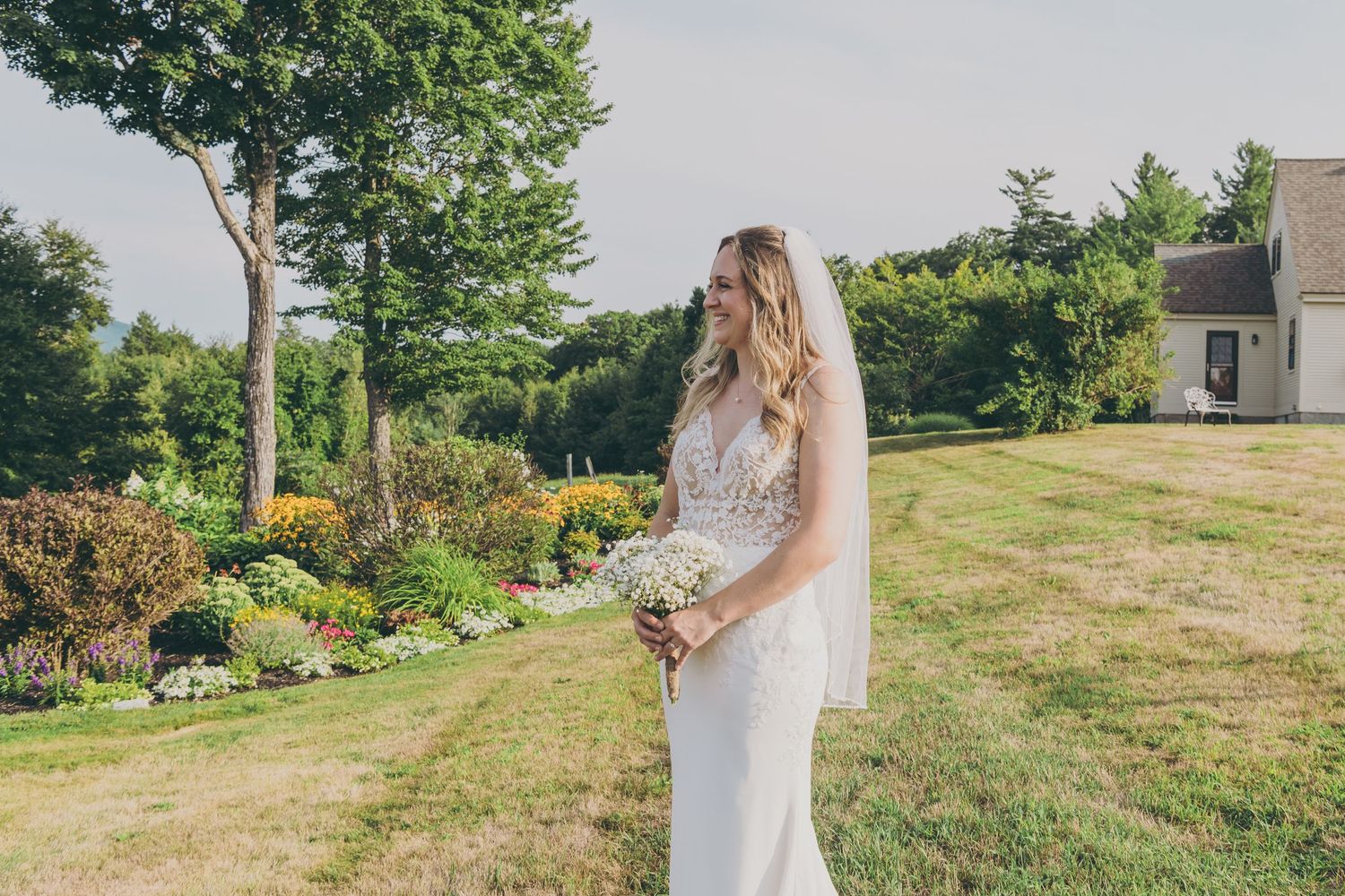 Bride in fitted lace gown poses in garden setting with flowering bushes and trees.