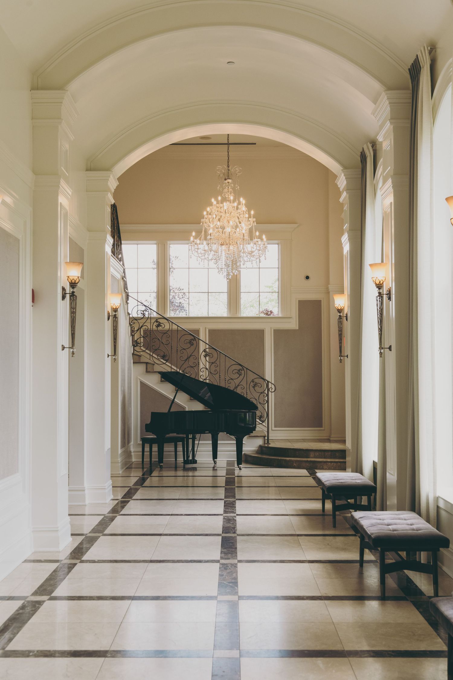 Elegant hallway with crystal chandelier, grand piano, tiled floor, and bench seating beneath white arched columns.