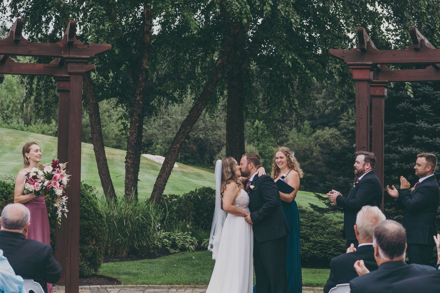 An outdoor wedding ceremony takes place under a wooden pergola surrounded by lush trees and greenery at Atkinson Country Club.