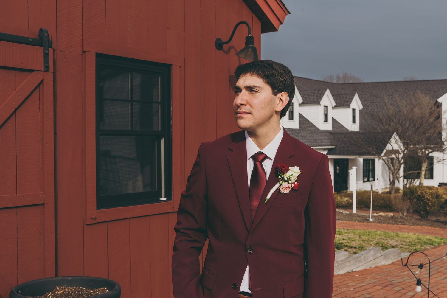 A person in a burgundy suit and tie stands in front of a red barn with white houses visible in the background.