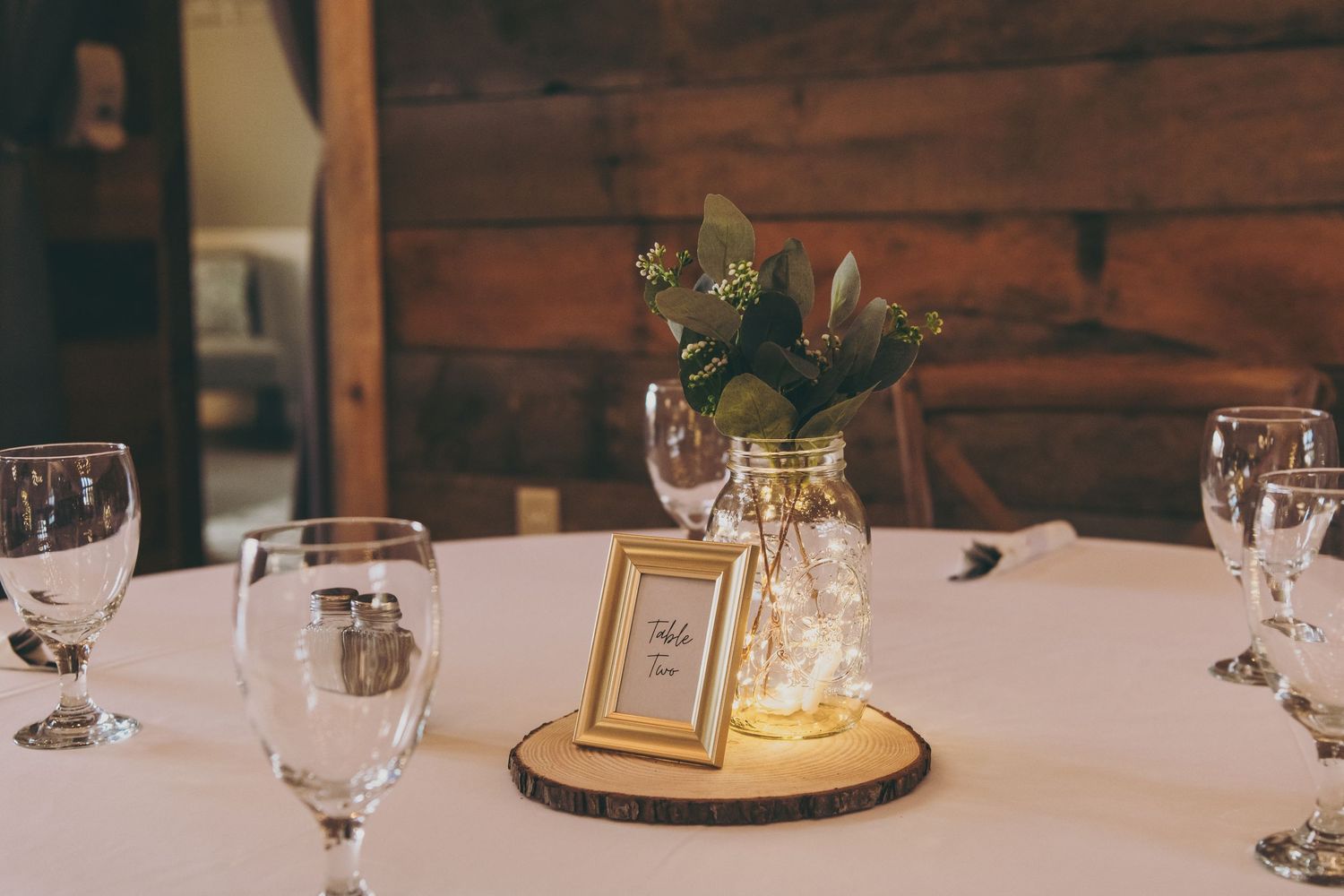 A rustic wedding table centerpiece with eucalyptus in a mason jar, table number, and wine glasses on a white tablecloth.