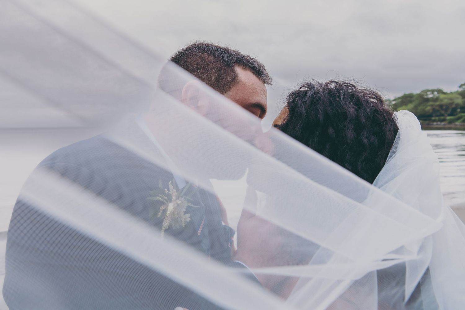 A romantic couple shares an intimate moment under flowing white sheer fabric outdoors near a body of water.