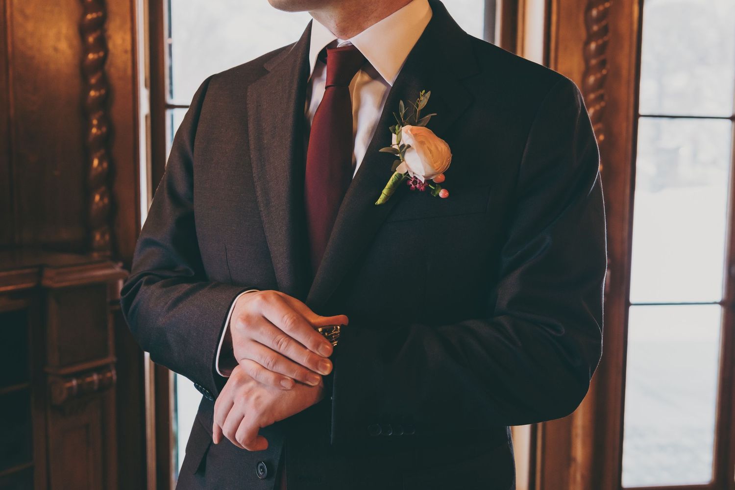 A groom in a classic black tuxedo adjusts his boutonniere before a wedding ceremony.