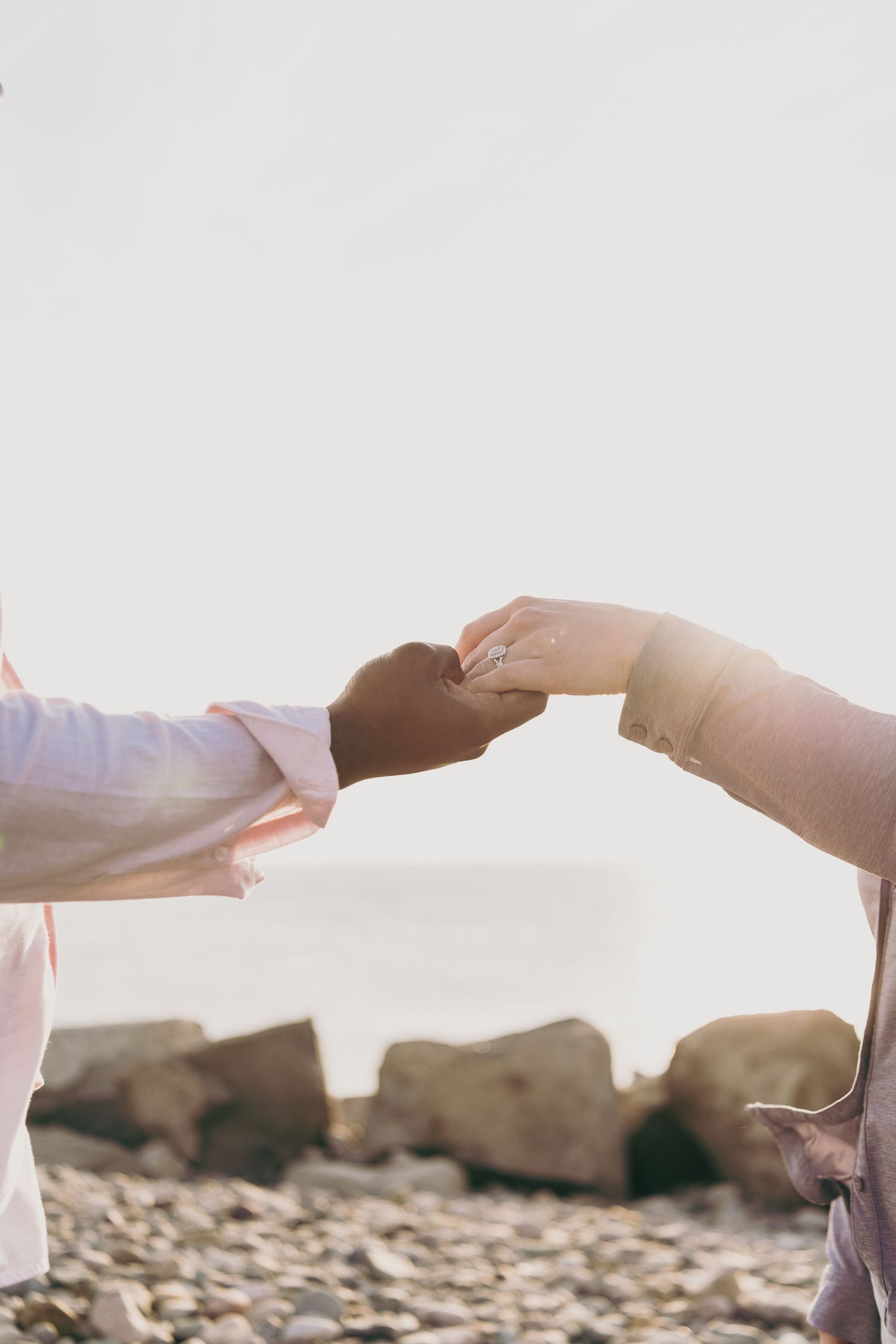 Two hands forming a heart shape silhouetted against a rocky beach background at sunset.