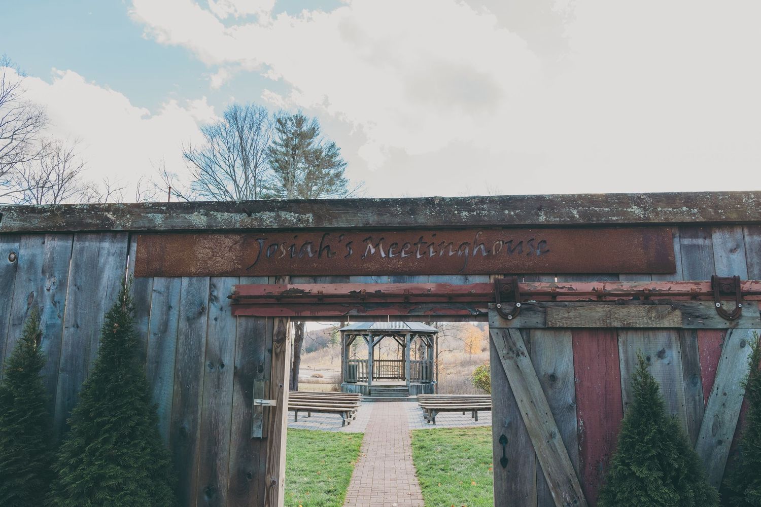 A rustic wooden archway leads down a stone path through evergreen trees to a distant building on a cloudy day.
