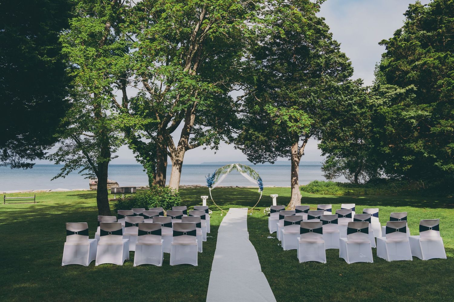 White chairs arranged in rows for an outdoor wedding ceremony with large trees and a cloudy sky in the background.