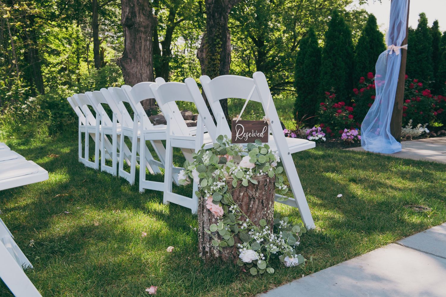 White ceremony chairs set up on grass with floral arrangements and fabric draping for an outdoor wedding.