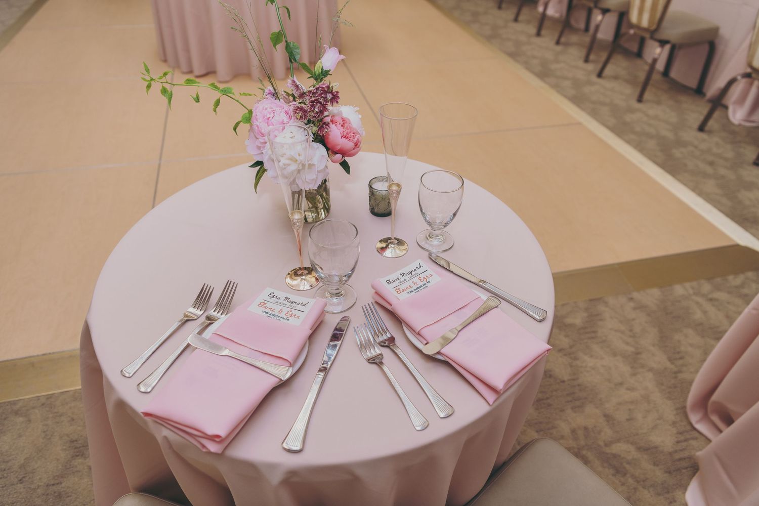 An elegant wedding table setting with pink napkins, floral centerpiece, and crystal glassware on a round ivory tablecloth.