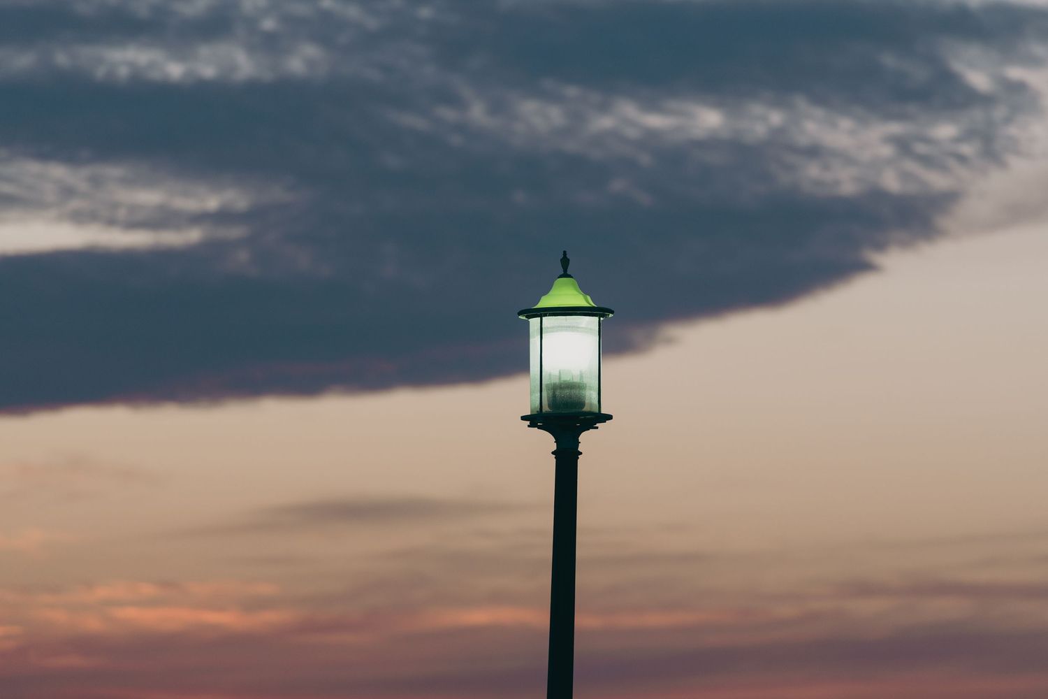 Black street lamp with green-tinted light glowing against a dramatic sunset sky with dark clouds. Location: Scituate Lighthouse