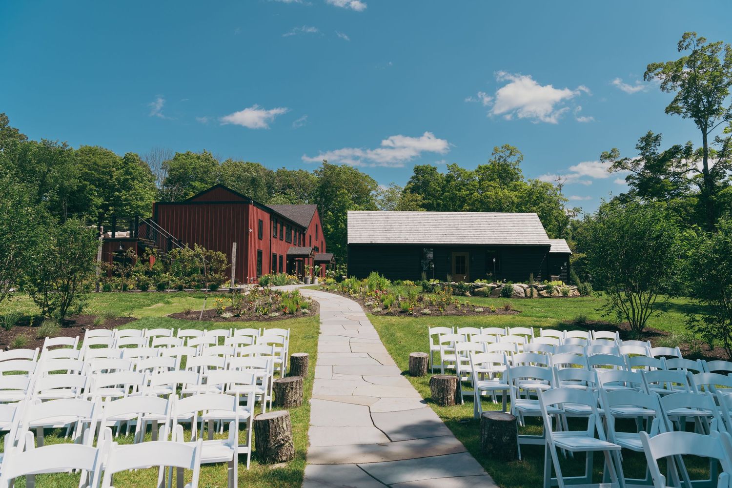 White chairs arranged for an outdoor wedding ceremony with a red barn and white building in the background on a sunny day.