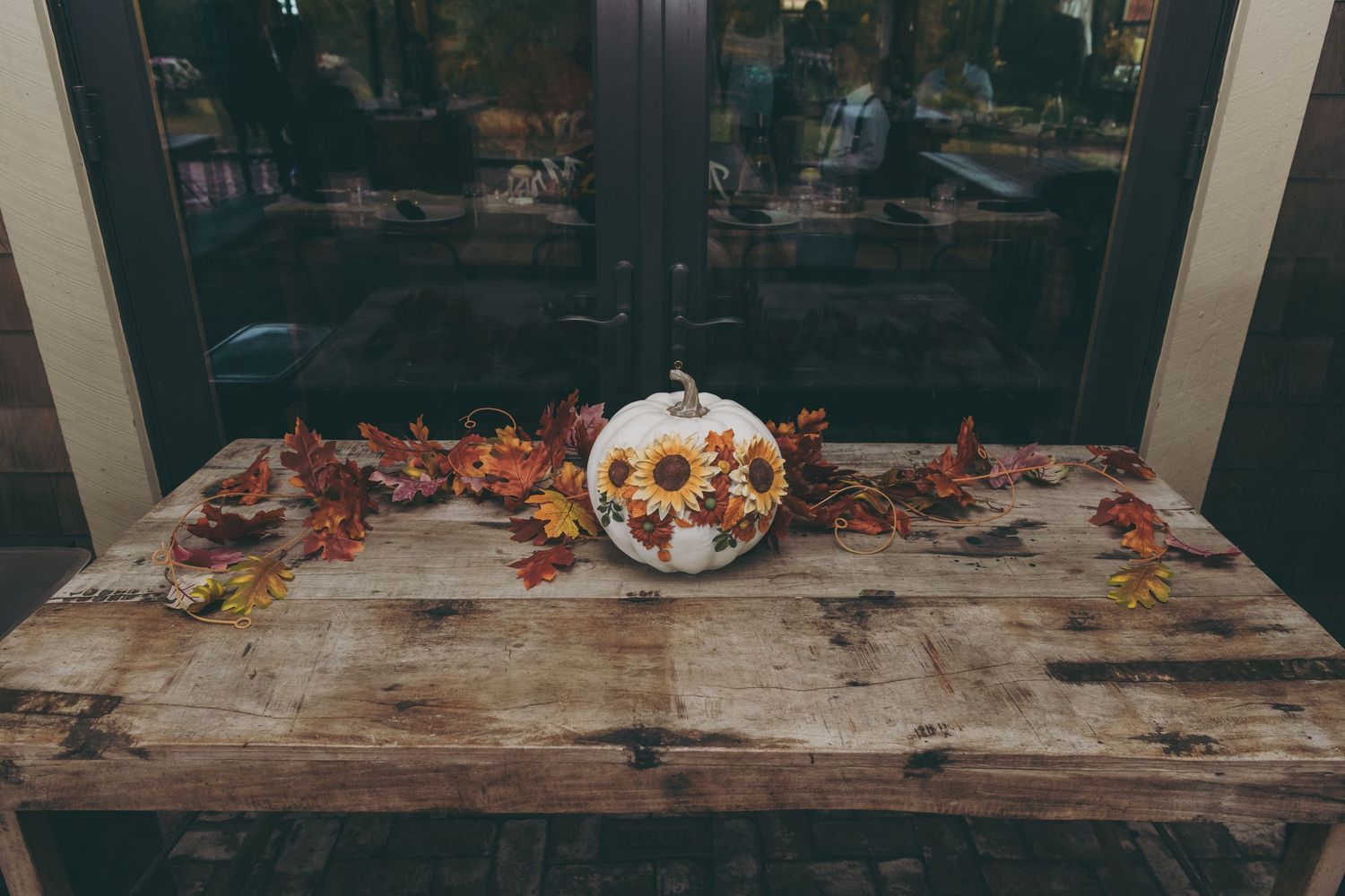 Rustic wooden table decorated with autumn leaves and a pumpkin centerpiece by a window.