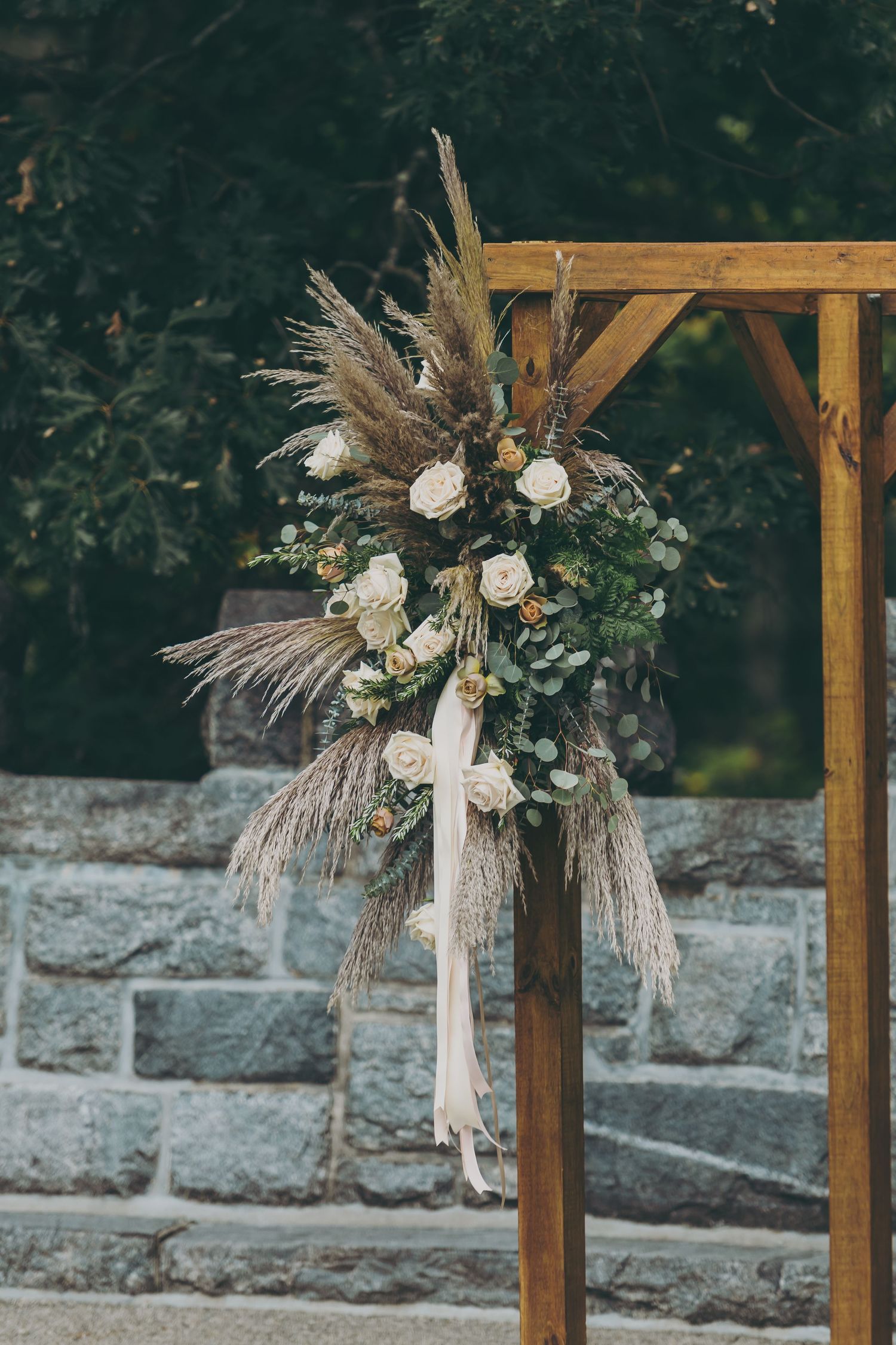 Wooden ceremony arch decorated with dried pampas grass and white flowers against a stone wall backdrop.