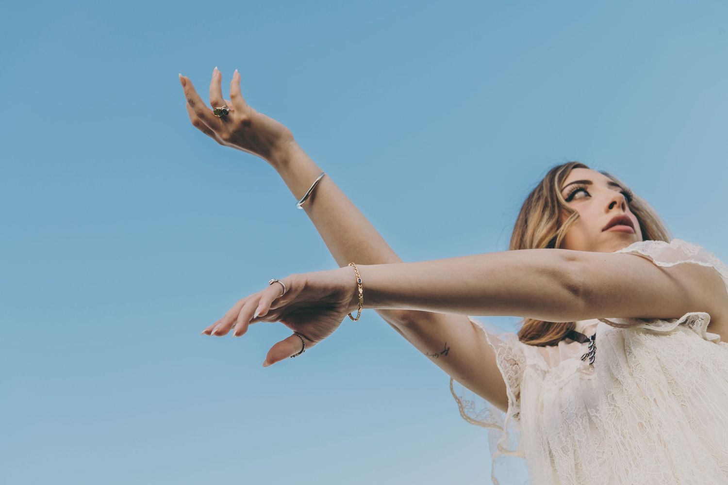 Person in white dress raises arms joyfully against blue sky background.