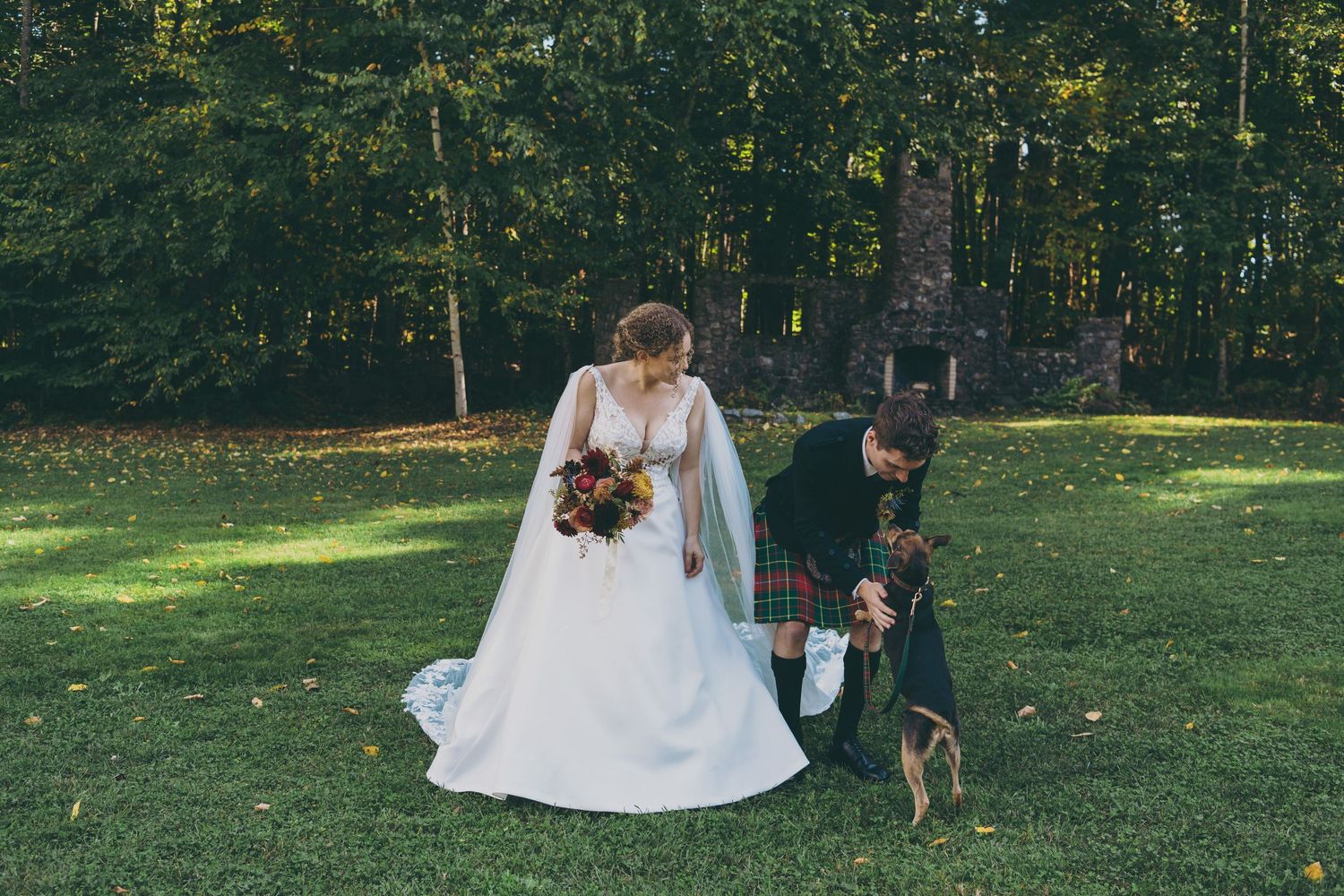 Bride in flowing gown walks with small dog in elegant garden setting.